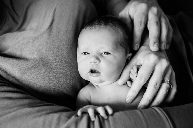 A natural and candid black and white baby photo showing the babys hands and the parents hands. The baby is the centre of the image and is being cuddled in a candid way by their parents. This is a documentary newborn photo.