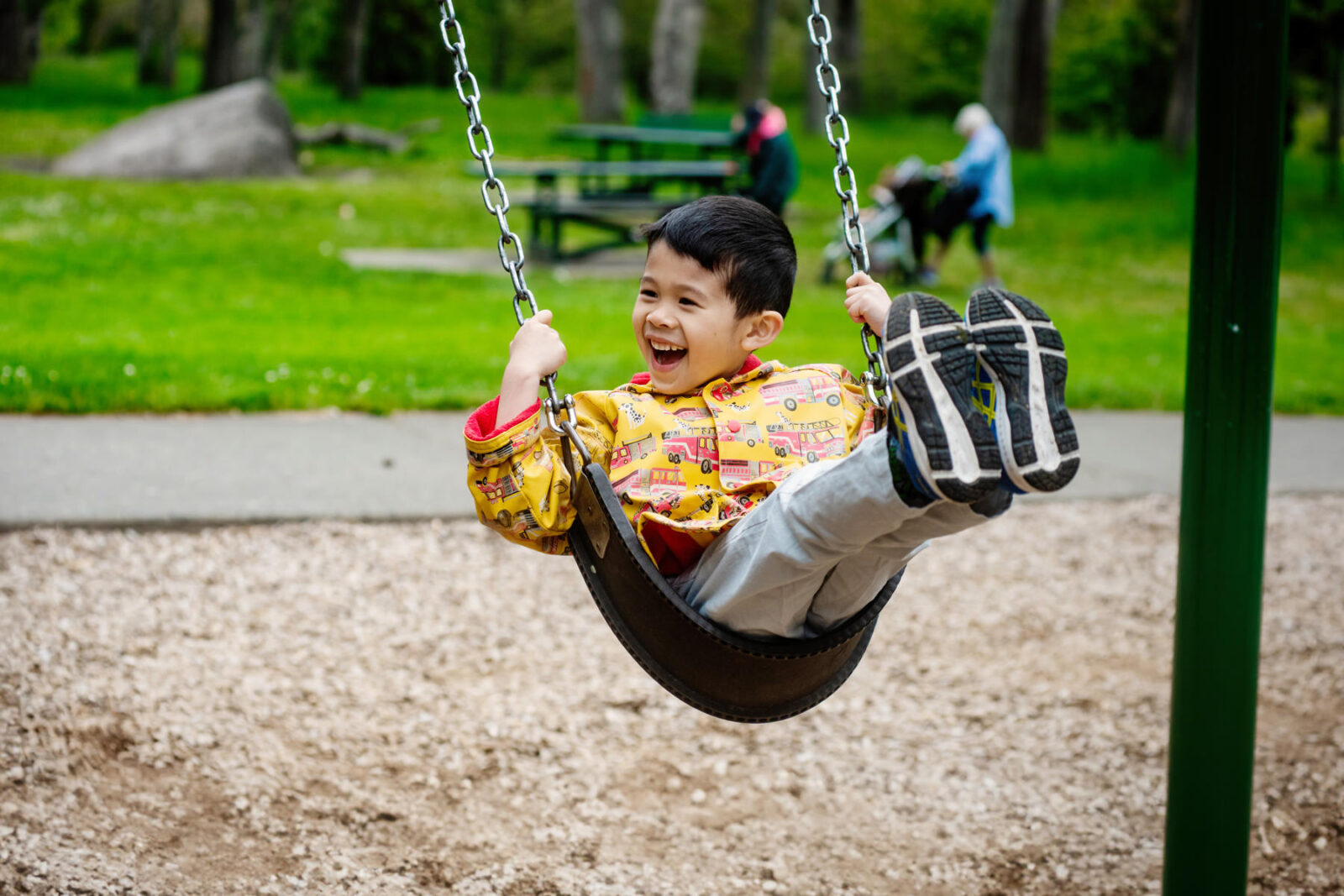 A happy child wearing a yellow jacket is smiling as he plays on the swings at the Beacon Hill Park play area.