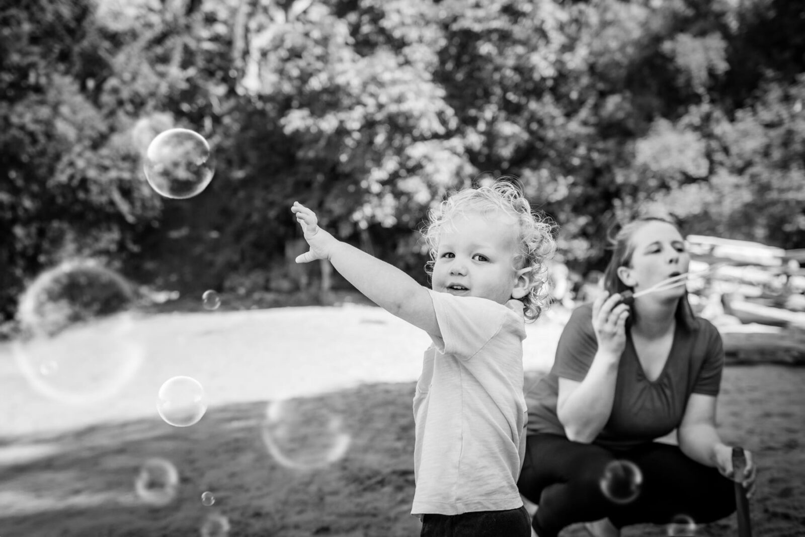 A blonde two year old boy pokes at bubbles as he plays with his mom in the sand at Hollydene beach in Victoria Bc