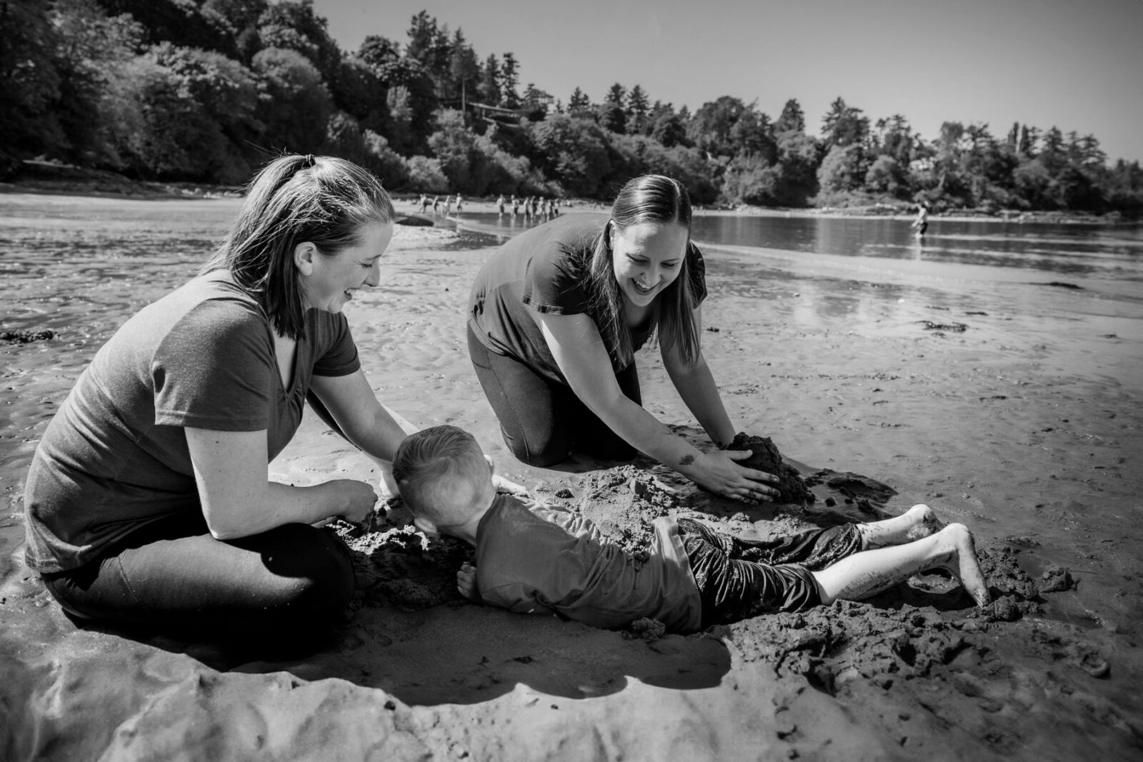 Candid beach photos of a staycation in Victoria BC at Hollydene park. A young boy is getting buried in sand by his moms. Photographed in black and white.