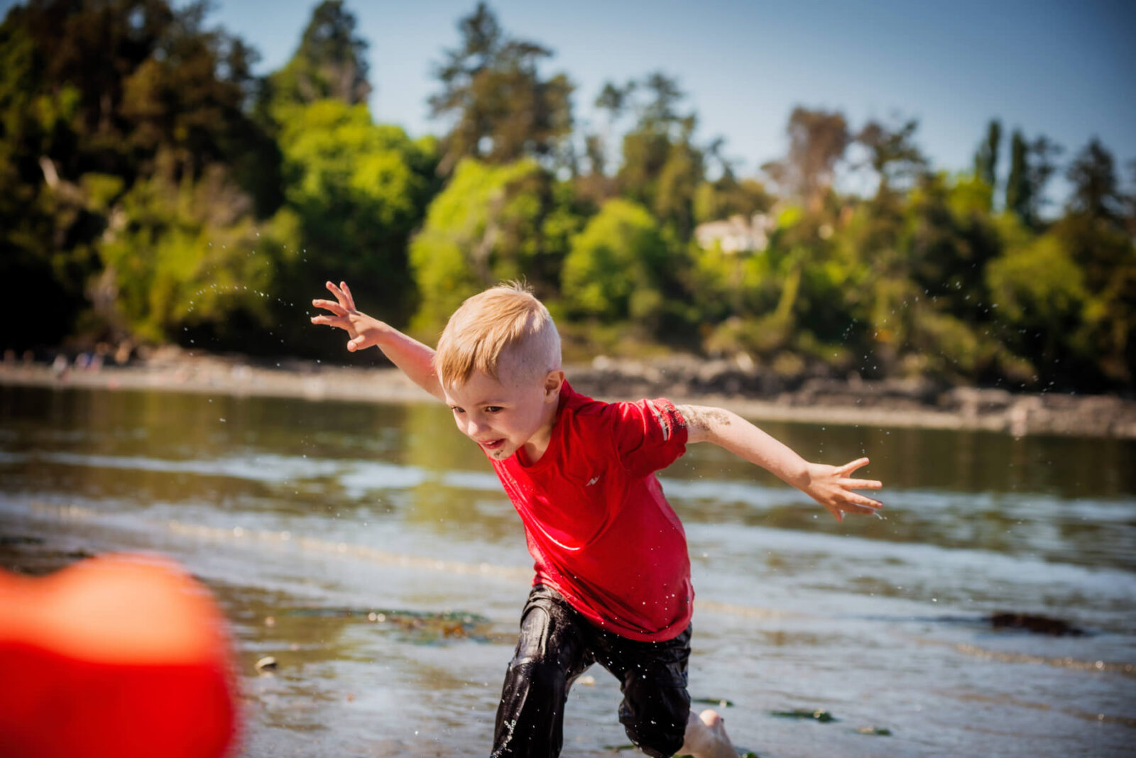 A color photo of a blonde boy in a red shirt playing in the dirt mud and water at Holydene beach in Victoria BC