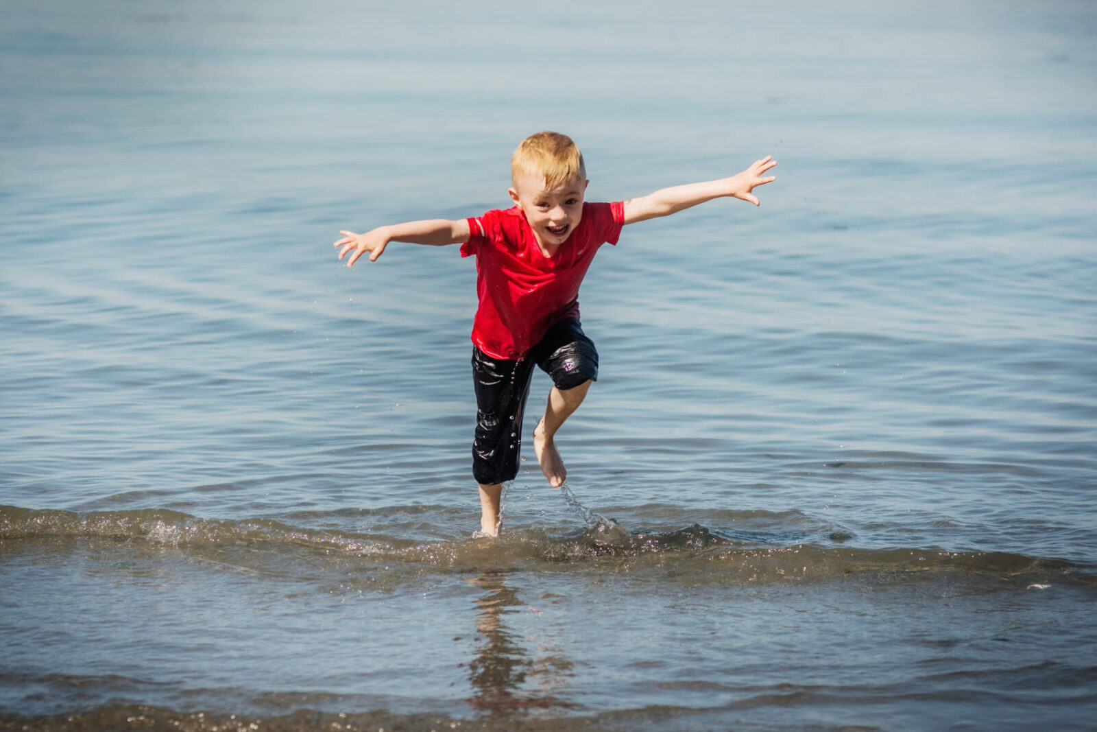 A color photo of a blonde boy in a red shirt playing in the dirt mud and water at Holydene beach in Victoria BC