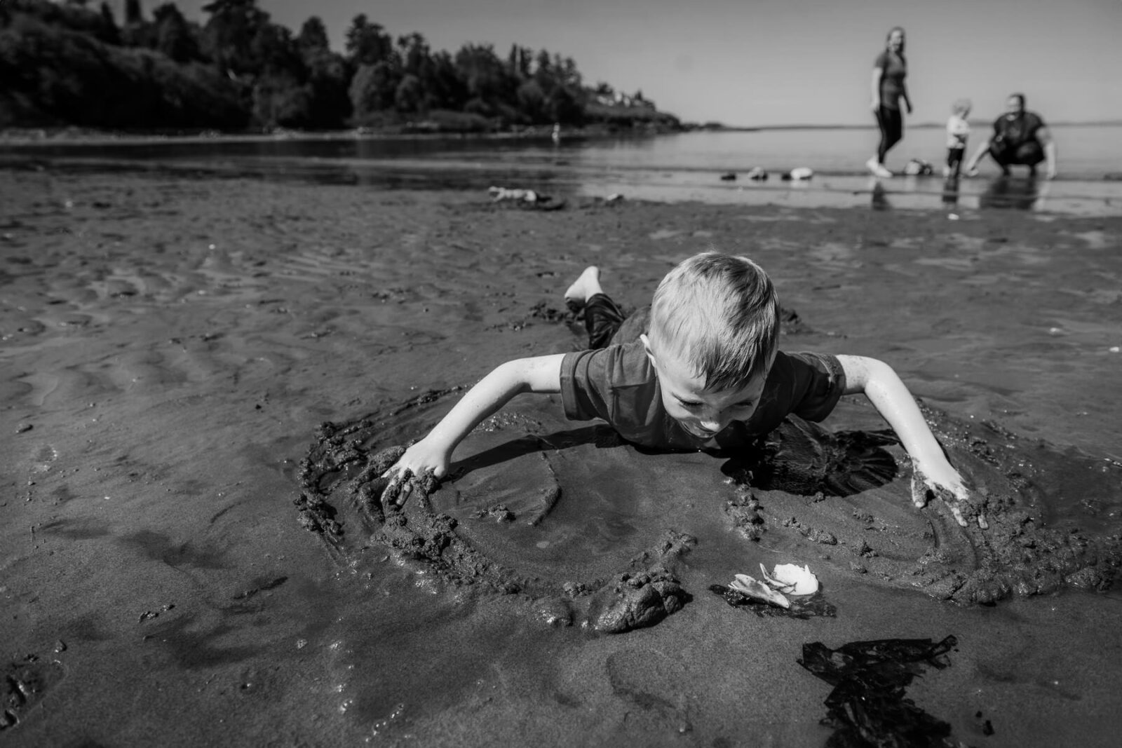 A black and white documentary photograph of a young boy playing in the mud on the beach in Victoria BC during a family photo session.