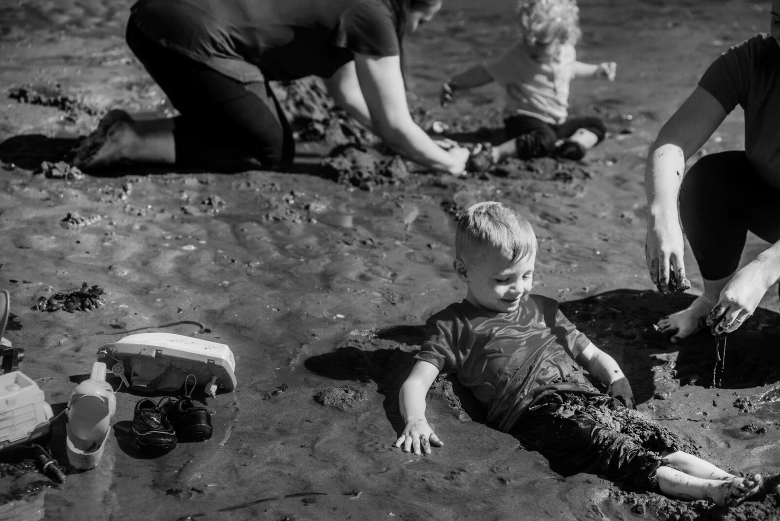 A black and white photo of a boy getting buried in sand by his parents at the beach in Victoria BC. This is a documentary family portrait by a professional photographer.