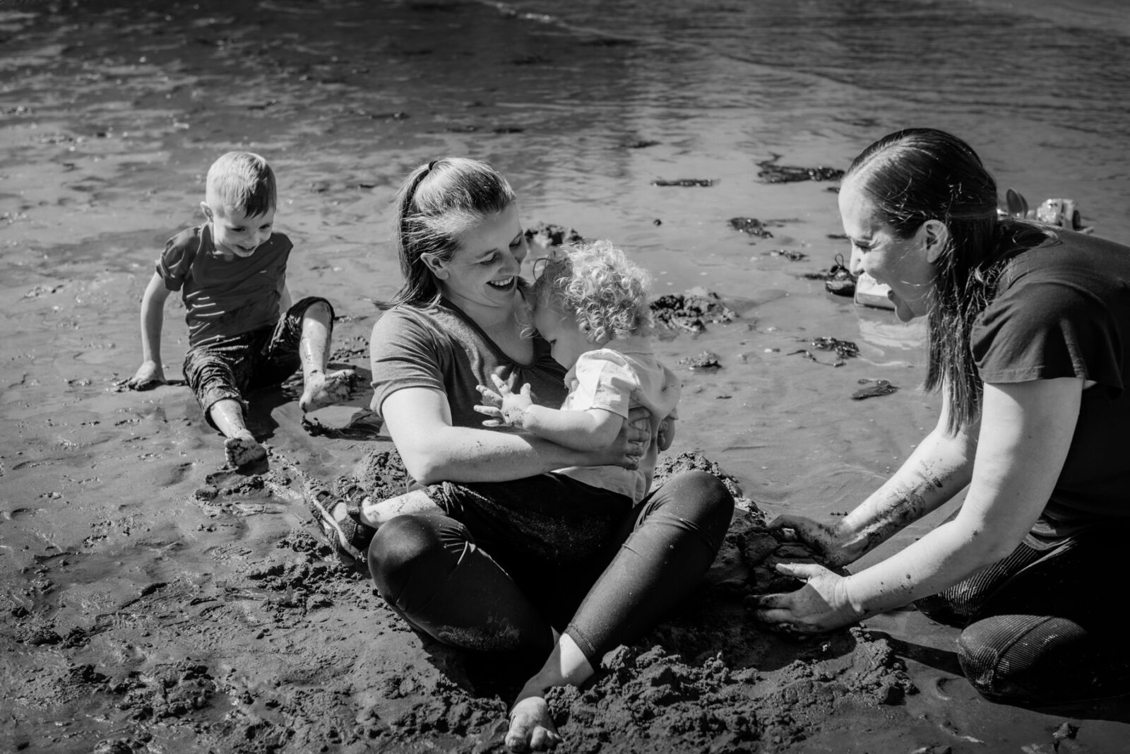 A documentary family portrait of two moms with their sons playing on the beach in the mud and getting wet. The beach is Hollydene park in Victoria BC.