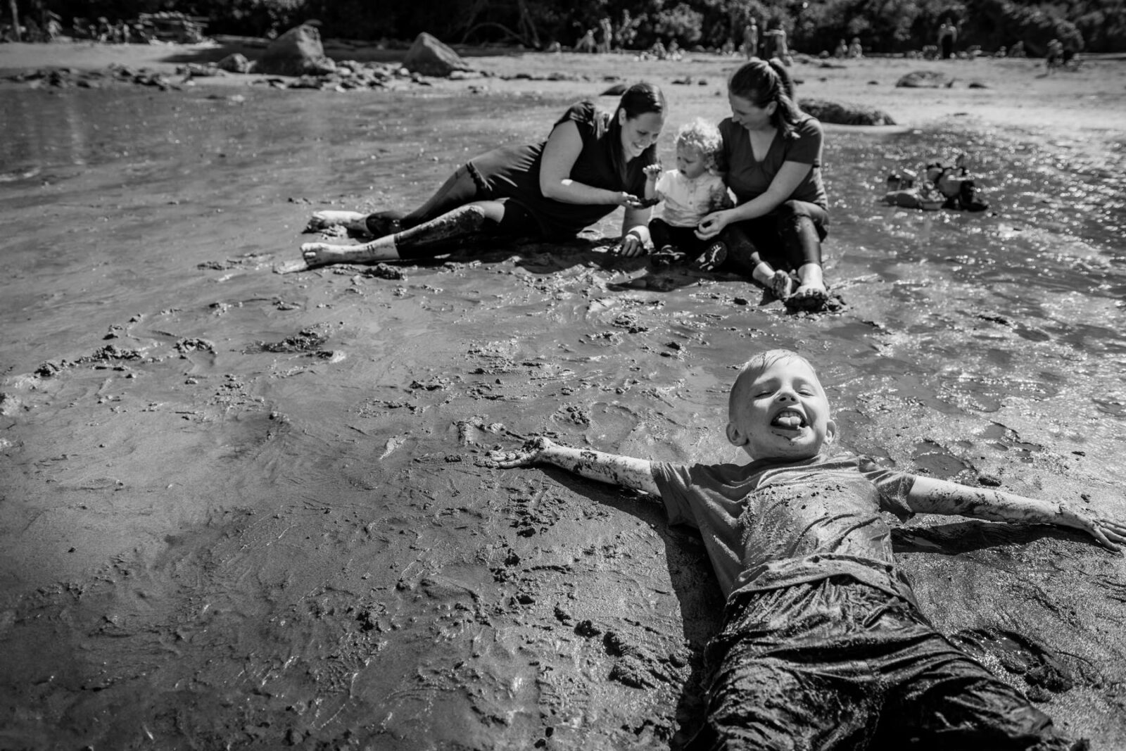 A funny photo in black and white taken at Hollydene Beach in Victoria BC. A boy has his tongue out as he makes a sand angel in the mud while his moms and brother play with shells behind him.