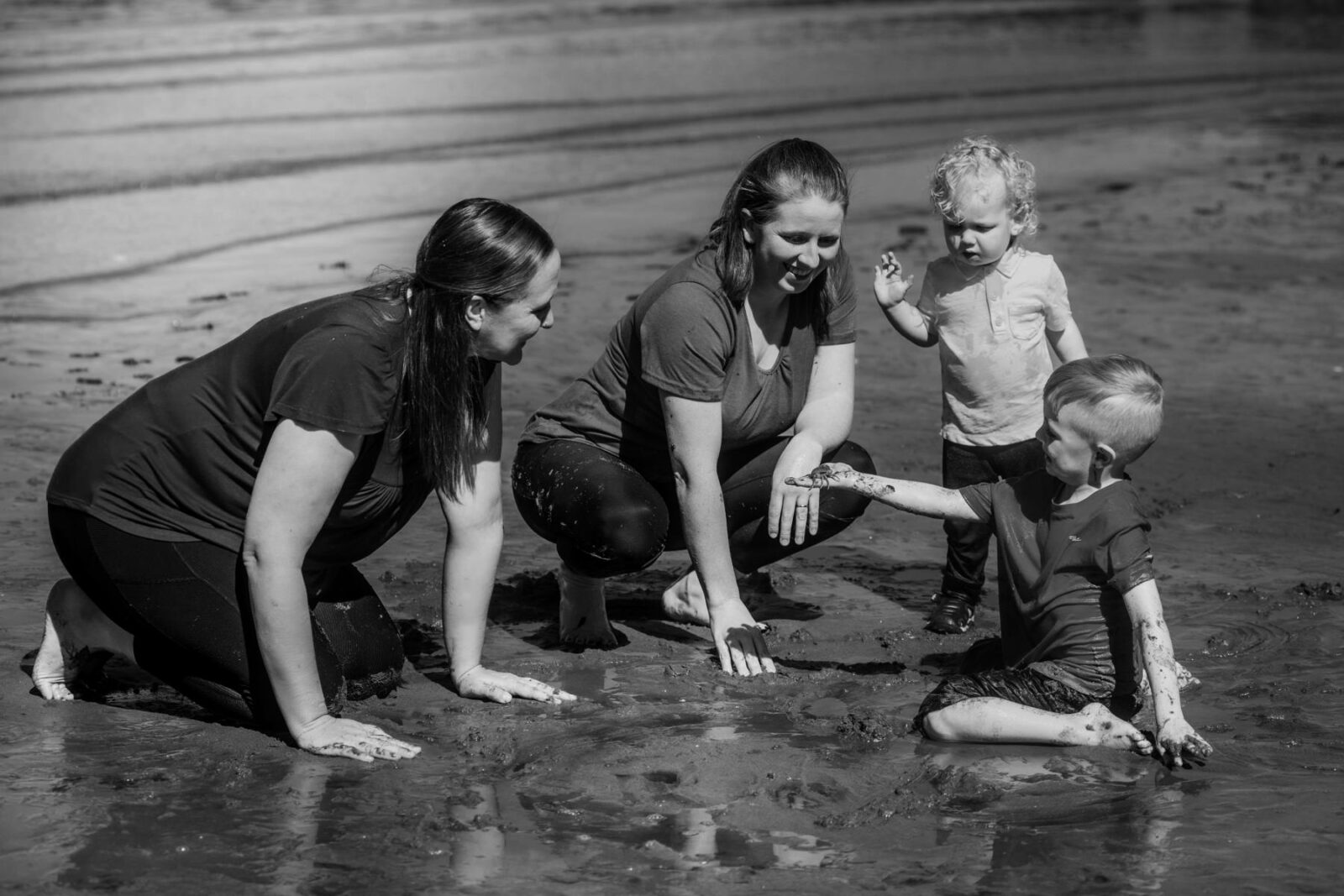 A candid moment capture in Black and White of a family playing at Hollydene Beach in the mud. A young boy shows his mom a crab.