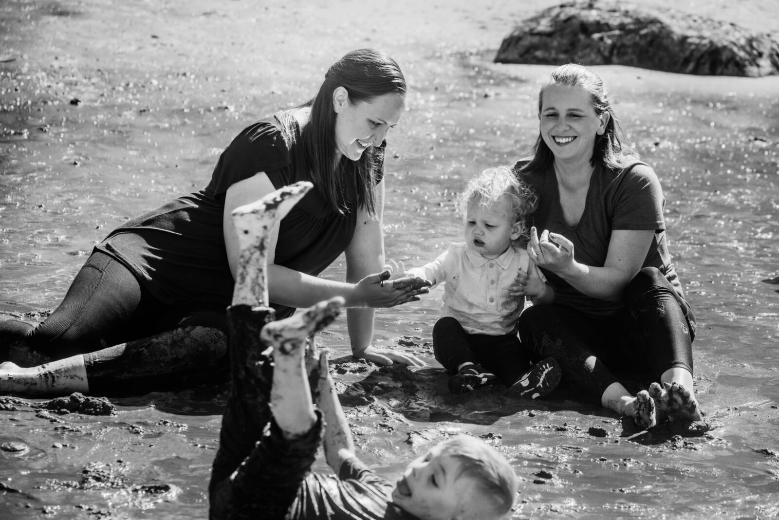 A silly photo of a blonde boy rolling in the sand while his moms and brother are playing in the mud behind him. This photo is in black and white and was taken at Hollydene park in Victoria BC