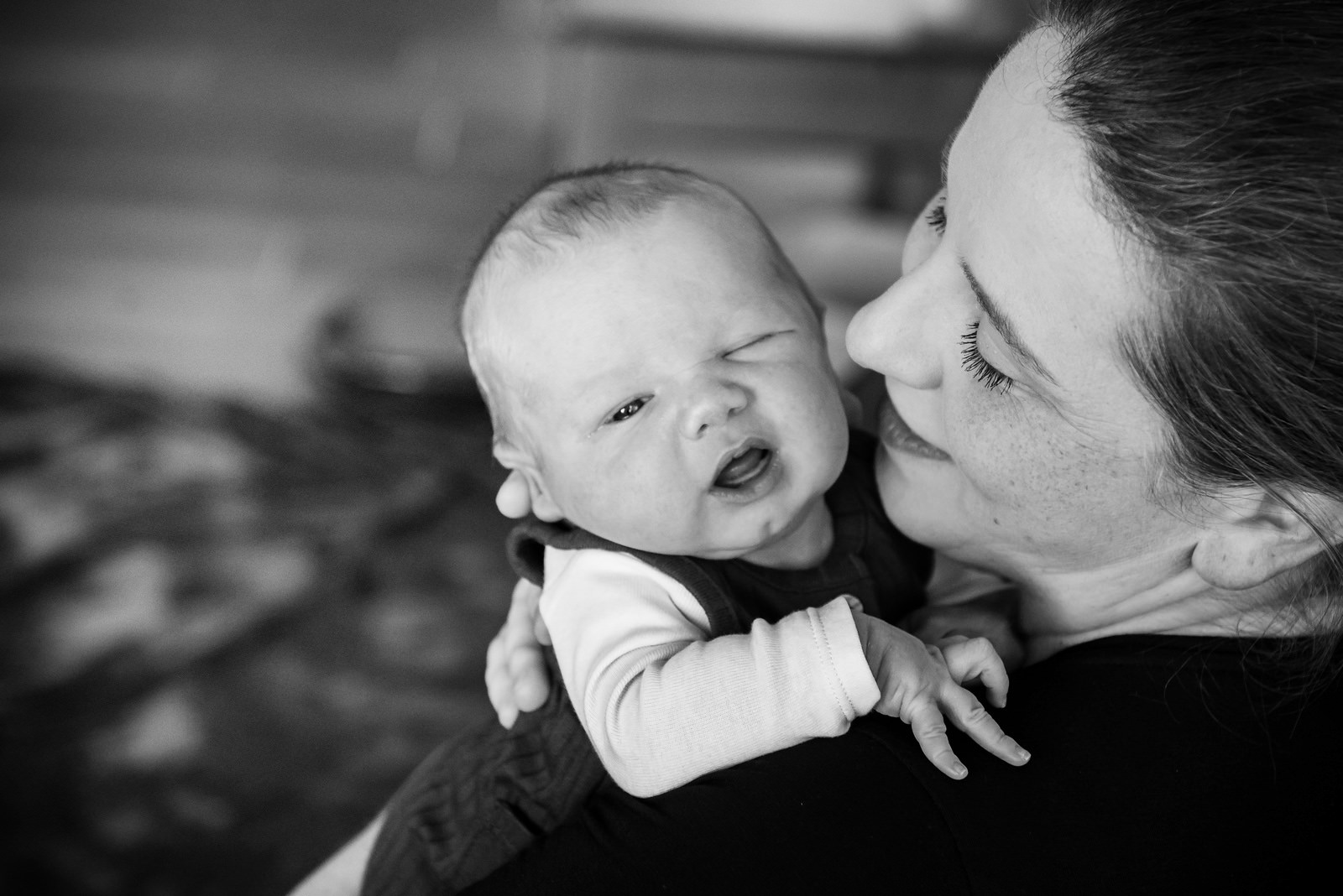 A black and white professional newborn portrait of a baby winking and looking cute as his mother holds him. Taken in Victoria BC