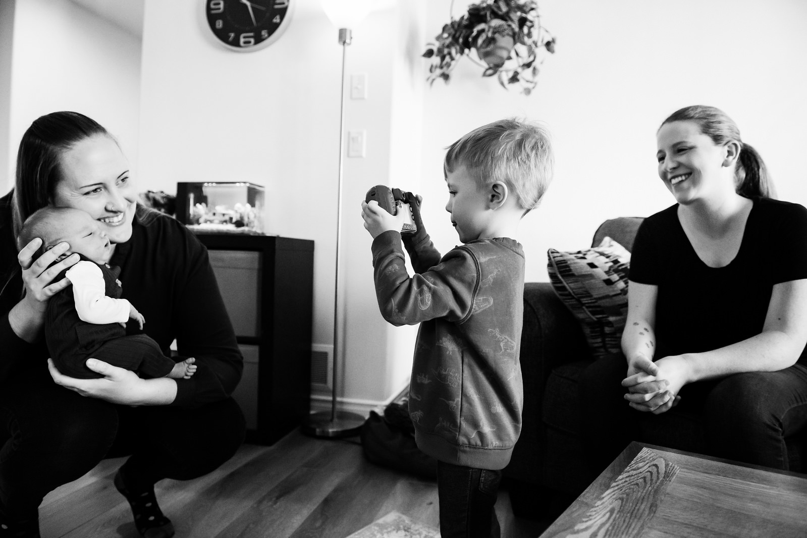 A candid moment where a new big brother is photographing his newborn sibling with his moms laughing. This is an at-home newborn photography session taken in Victoria BC