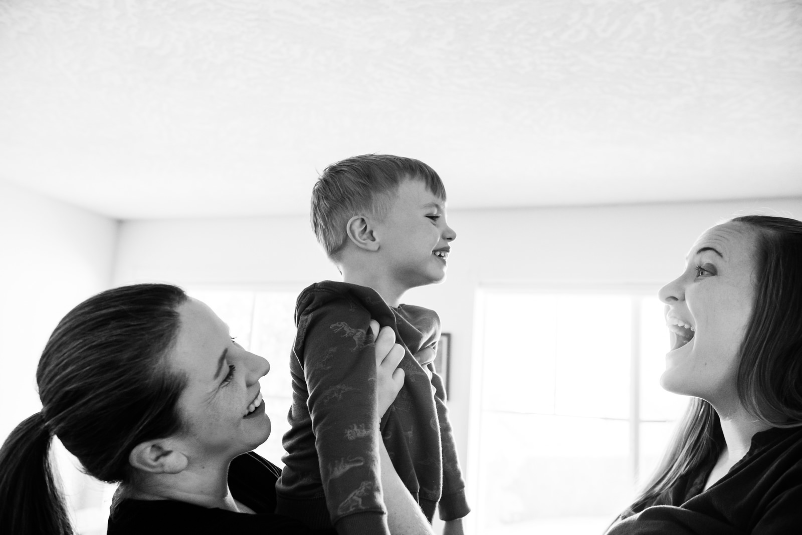 Two moms holding their son up in the air and making smiley faces at him in natural light in the living room of their home in Victoria BC