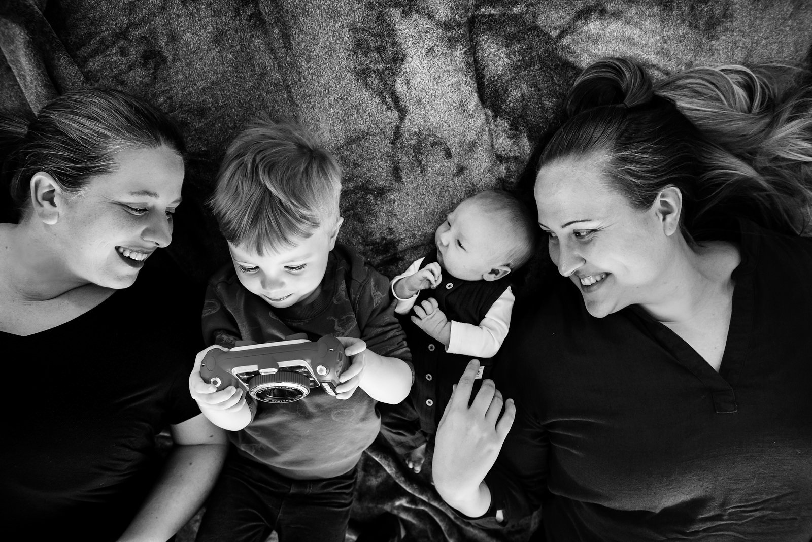 A black and white family portrait of a newborn baby, a toddler and two moms, laying on the floor looking at photos taken on their son's toy camera. They are on their living room floor in Victoria BC