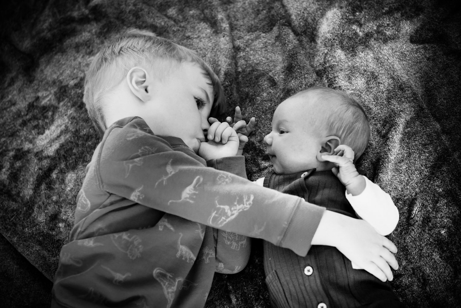 A toddler hugs his newborn brother while laying on the floor on a blanket in the living room in Victoria BC