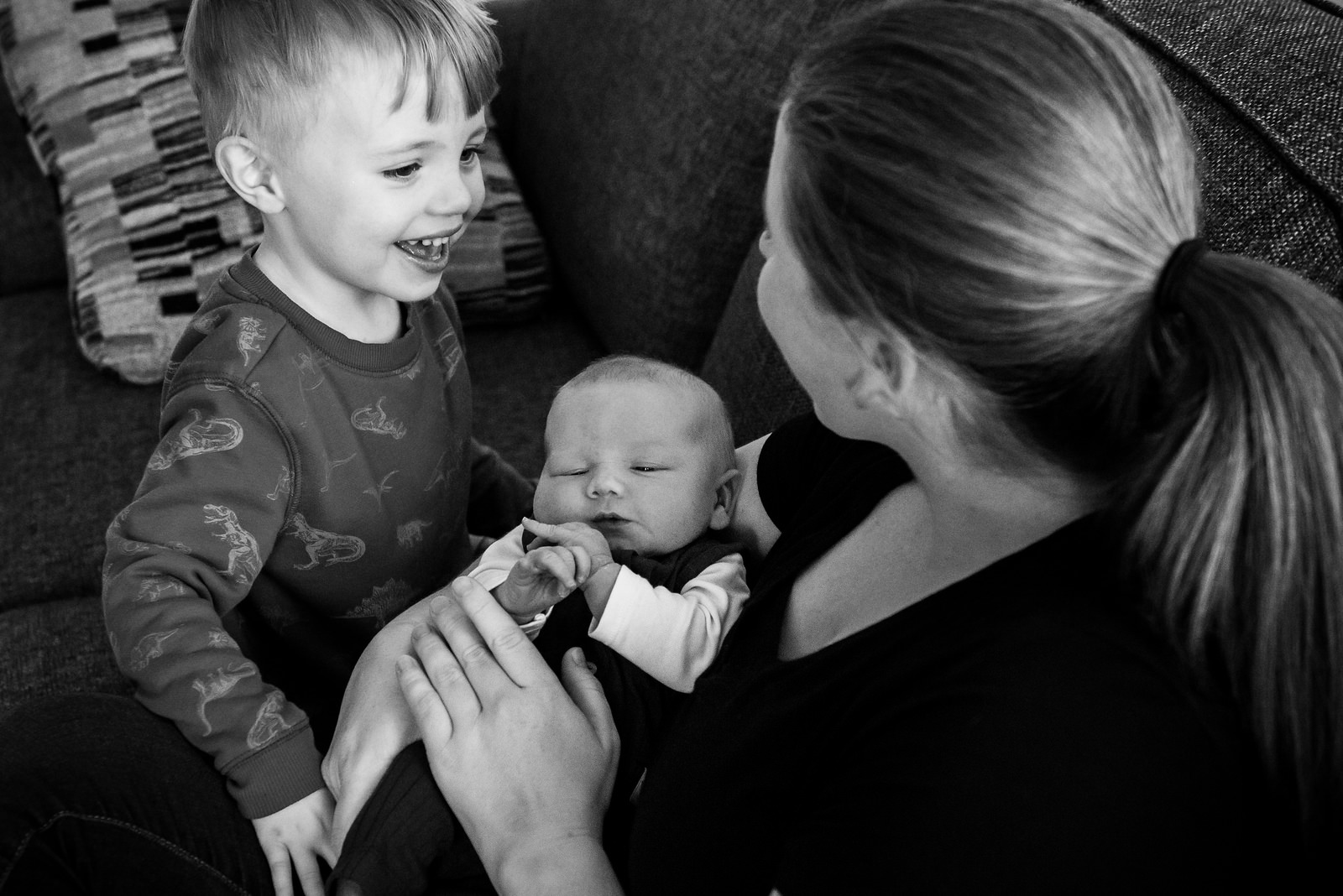 A toddler is smiling and laughing while his mother holds his newborn baby brother in their living room in Victoria BC