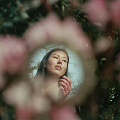 A creative fashion photo taken on 35mm analog film with a model looking off in the distance. her image is reflected in a mirror that is placed among some pink flowers at Abkhazi Garden in Victoria BC