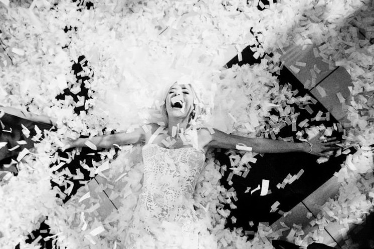 A bride surrounded by confetti laying on the floor with an expression of pure celebration. Photo taken in black and white in Victoria BC