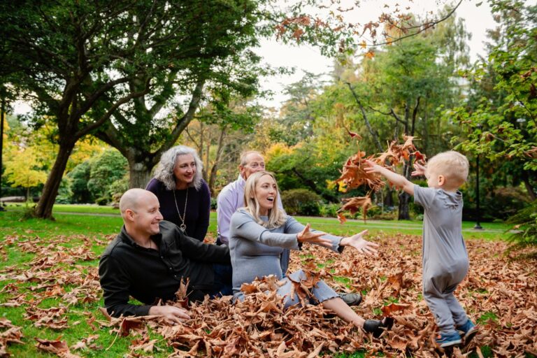 During a family mini session at Beacon Hill park a 2 year old grabs and tosses fall leaves at his parents and grandparents with everyone having happy expressions.