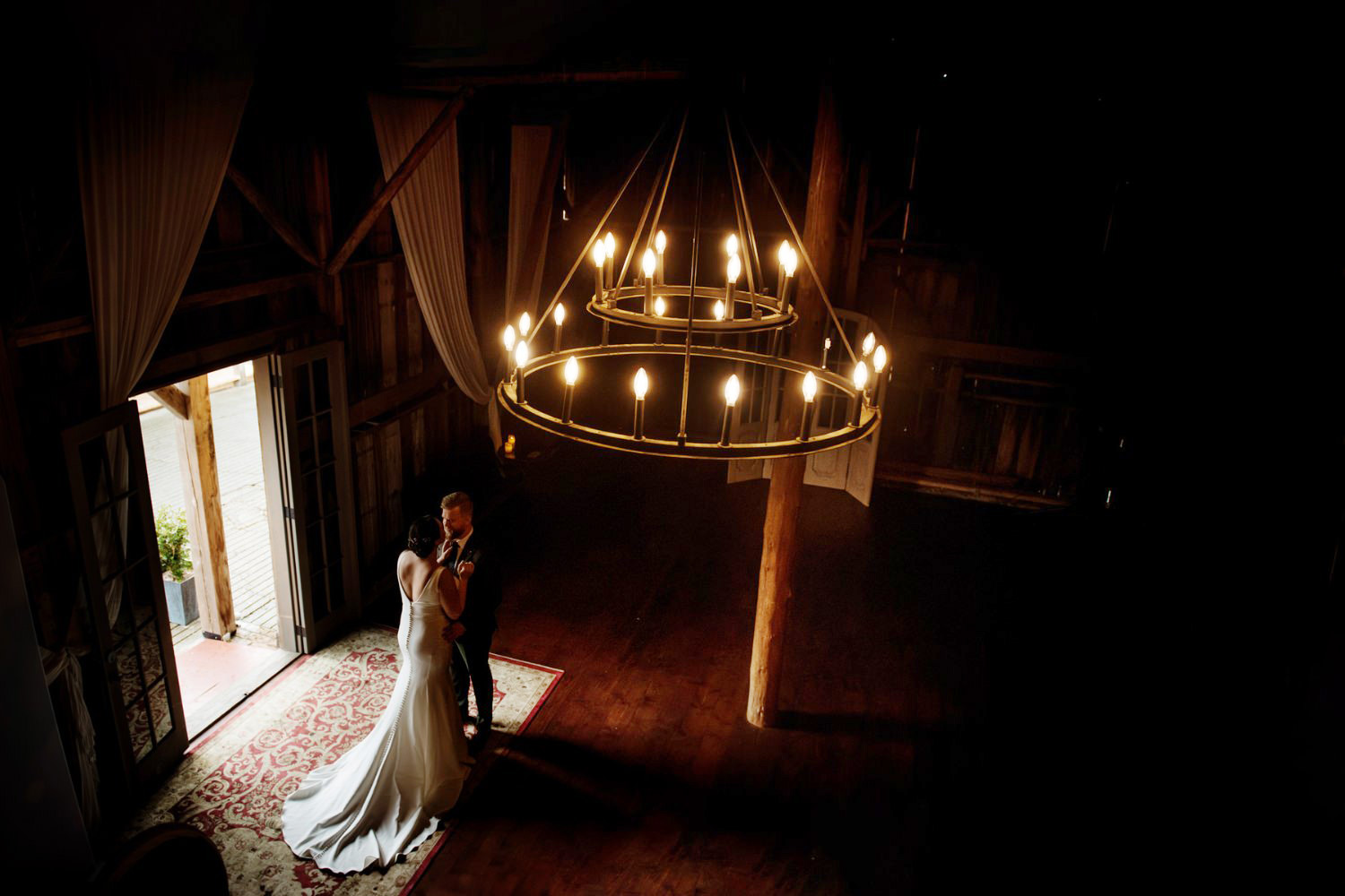 A couple stand in a barn on a farm at Langtry Walk Saanich near Victoria BC kissing under a candle chandalier in a rural barn.