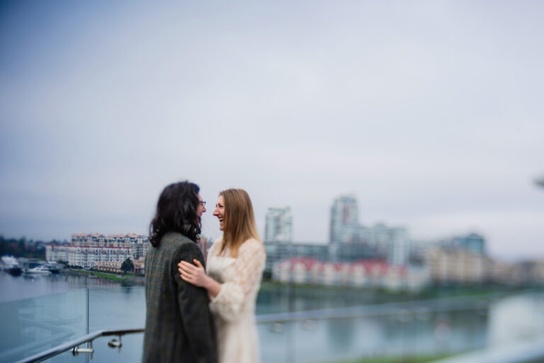 A lensbaby tilt-shift photo taken at the penthouse suite balcony at Inn at Laurel Point. A bride and groom look at each other while laughing and you can see Victoria in the distance.